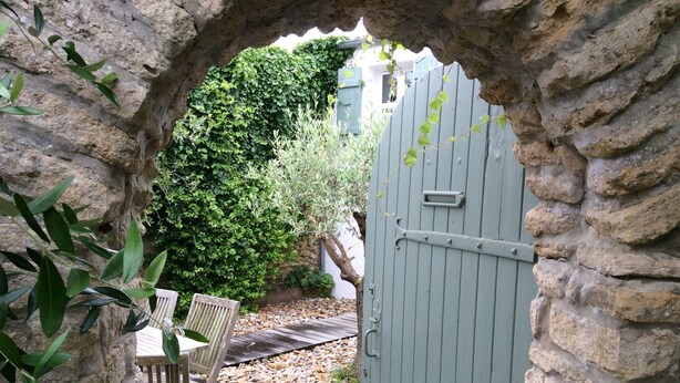 Un arco in pietra conduce a un pontile di legno con due sedie, un muro in pietra e un siepe verde sullo sfondo.