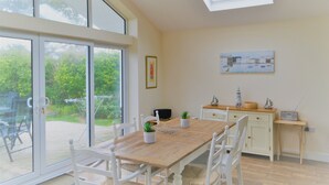 Un coin repas avec une table en bois, des chaises blanches, une porte vitrée donnant sur un jardin, un petit meuble avec une radio et un tableau accroché au mur.
