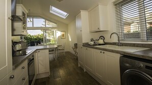 Une cuisine compacte avec des meubles blancs, un four encastré, un évier et une fenêtre équipée de stores.