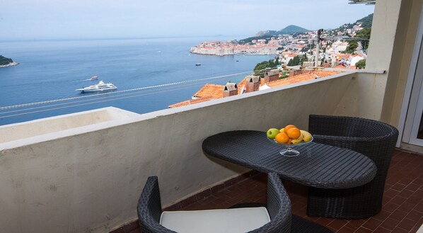 A balcony with wicker furniture, a fruit bowl, and a view of the sea and a historic town.
