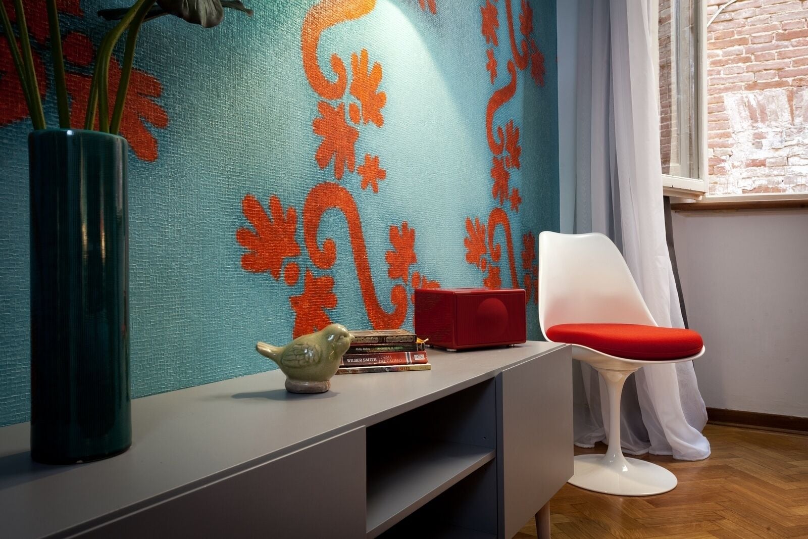 A modern room with a teal patterned wall, a white chair, a red cushion, a green vase, and a small bird figurine on a grey console table.