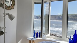 A bedroom with a large bed, a bedside table, a mirror, and a view of the ocean through a large window. - Runswick Bay Top Gallant Cottage - outside seating  - sea and beach views (Runswick Bay)