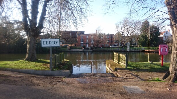 A sign reading "FERRY" near a body of water with a bridge, trees, and buildings in the background.