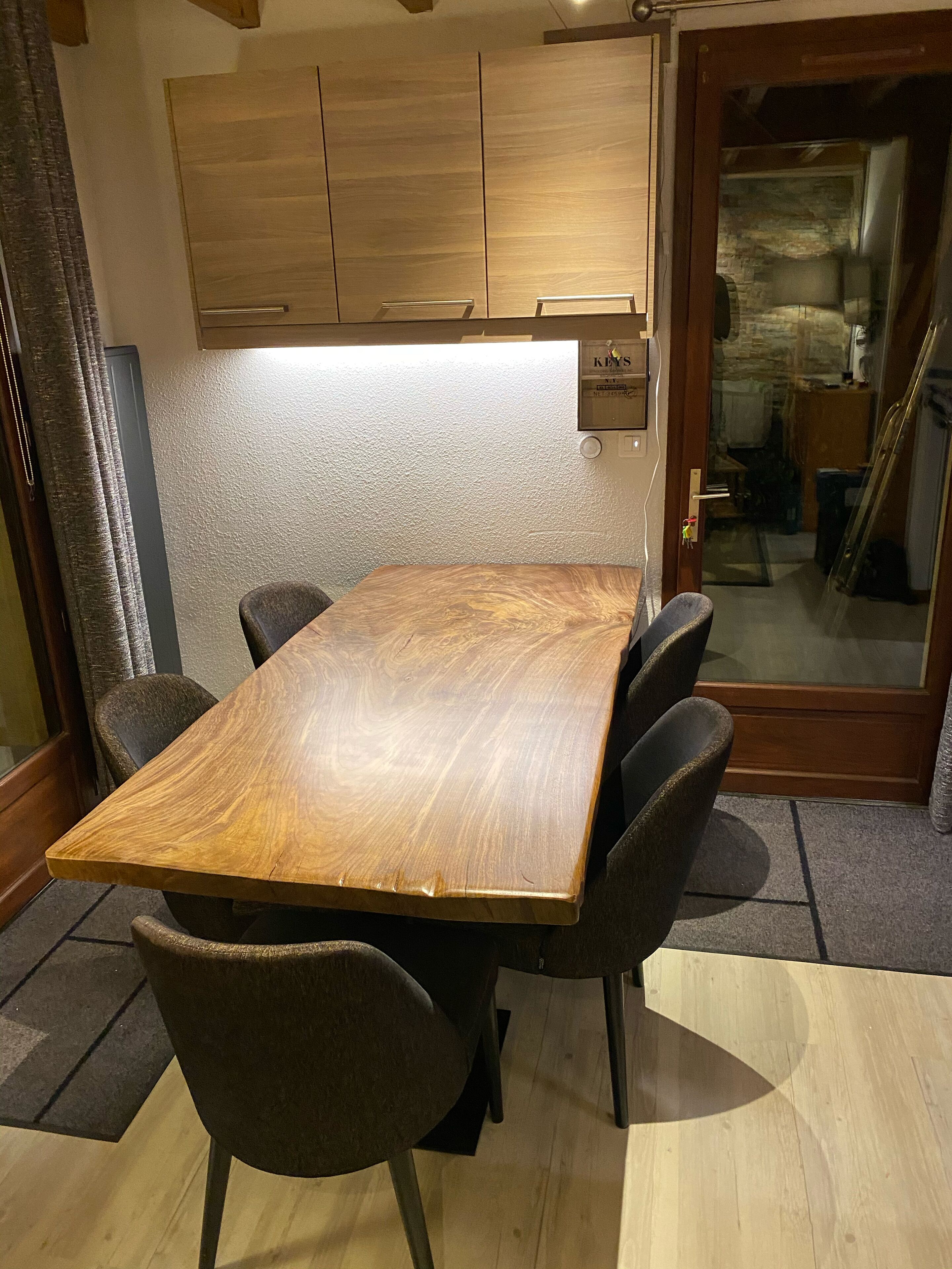 A wooden dining table with six chairs, a wooden cabinet above, and a glass door leading to a stone-walled room.