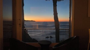 A balcony with a view of the ocean at sunset, featuring a railing and two wicker chairs.