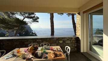 Un balcón con muro de piedra, sillas blancas, una mesa con frutero, bebidas y una tabla de madera.