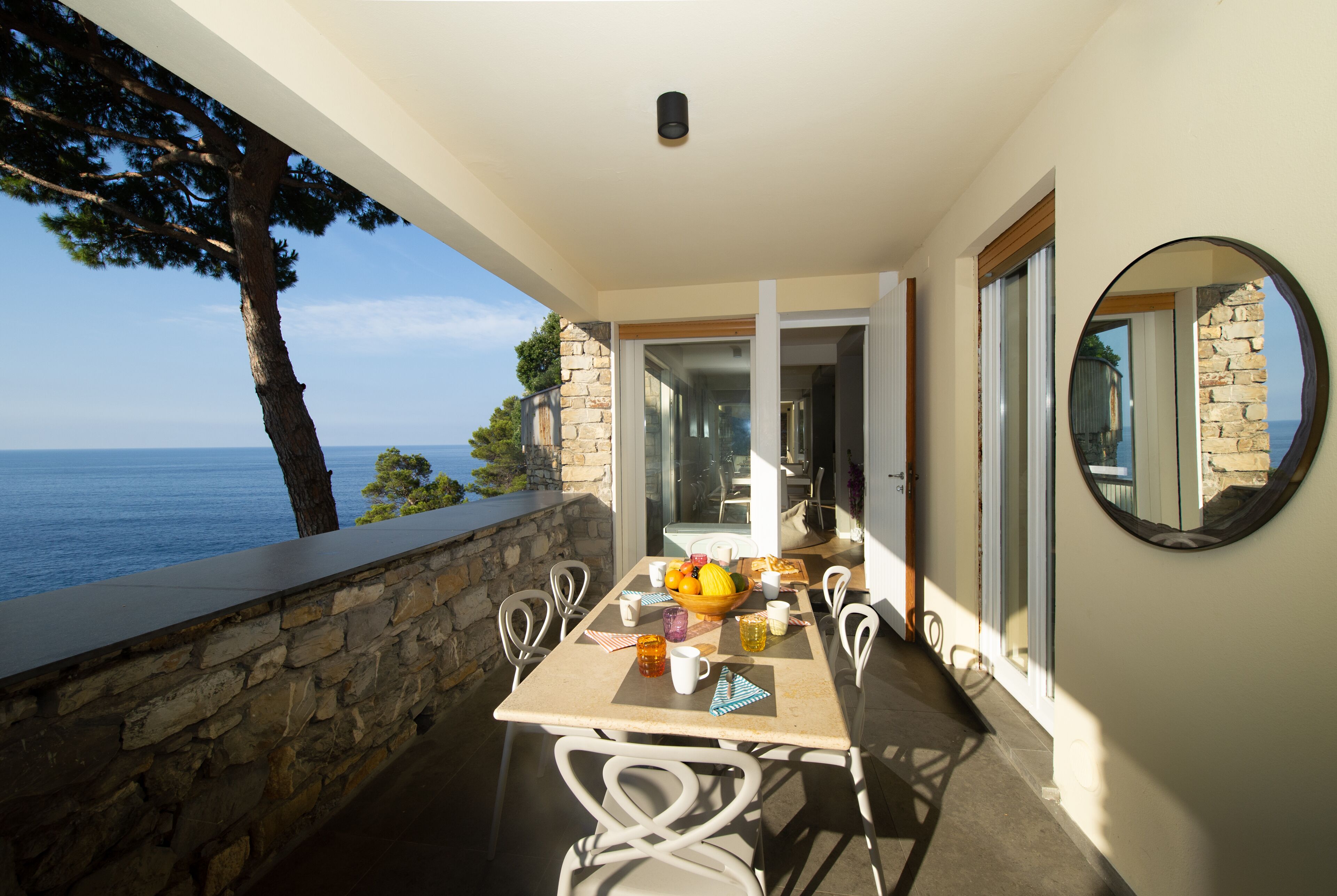 Un balcon avec un mur en pierre, une table avec des chaises, un saladier rempli de fruits et une vue sur la mer.
