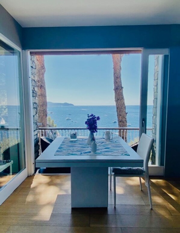 Un comedor con una mesa y sillas, un florero con flores, y vista al mar a través de amplios ventanales.