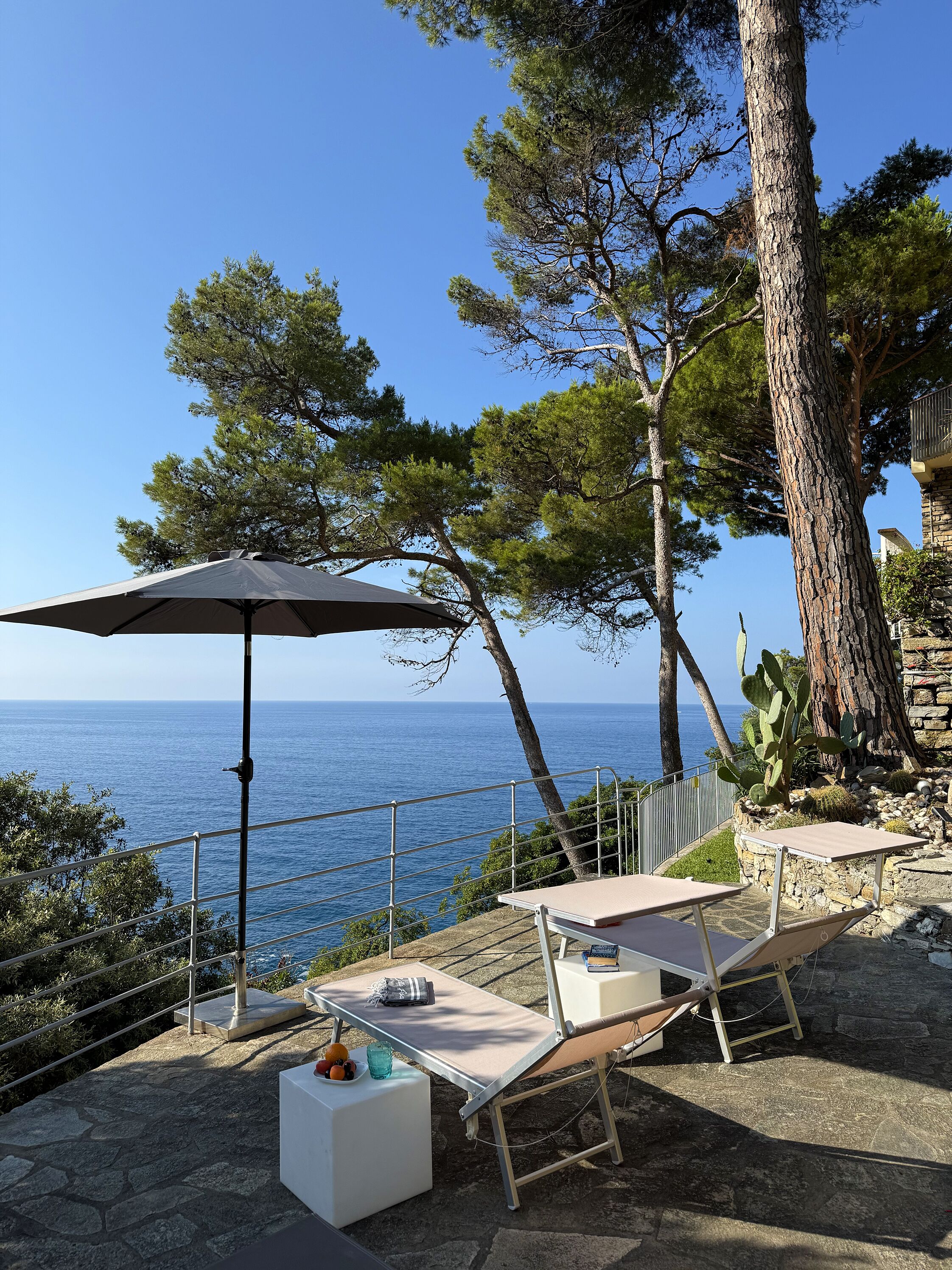 Una terraza con vista al mar, equipada con sillones de descanso y una mesa, rodeada de árboles y un muro de piedra.