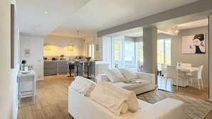 A spacious living area with a large white sofa, a dining table, and a kitchen in the background.