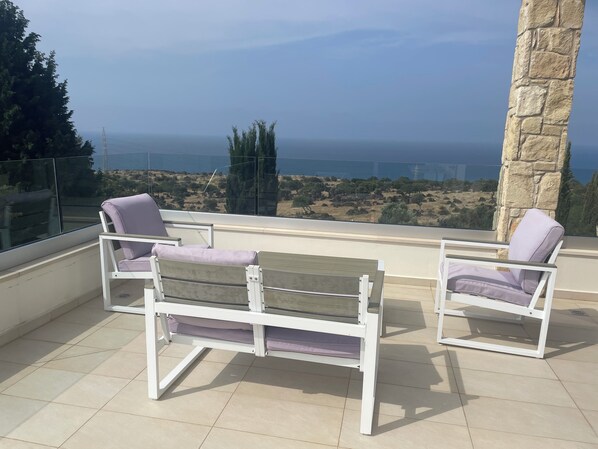 A balcony with two white chairs and a bench, overlooking a coastal landscape with trees and a clear view of the sea. - Villa Aphrodite Hills with Infinity Pool, Sea Views, Golf and Tennis (Kouklia)