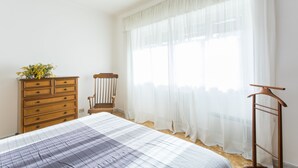 Un dormitorio con cama, cómoda de madera, una silla y ventana con cortinas de gasa.
