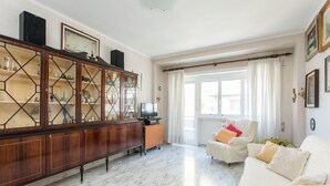 Una sala de estar con un sofá blanco, un mueble de madera con puertas de cristal, un televisor y una ventana con cortinas.