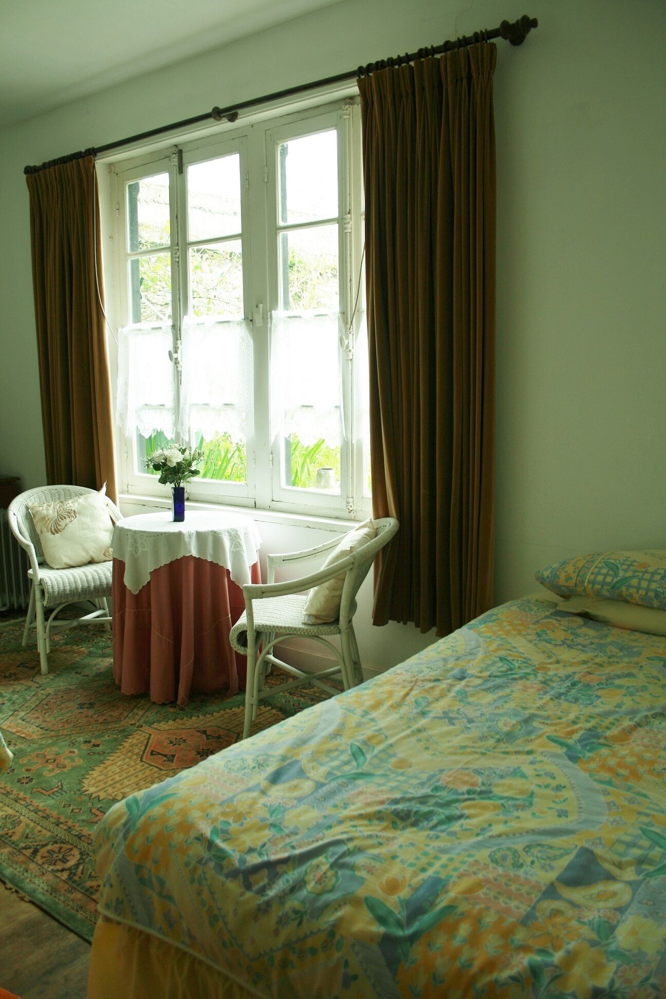 A room with a bed, a chair, a table with a vase, and a window with curtains.