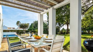 En udendørs terrasse med et glasbord dækket til spisning, omgivet af hvide stole, en grill og en swimmingpool i baggrunden.