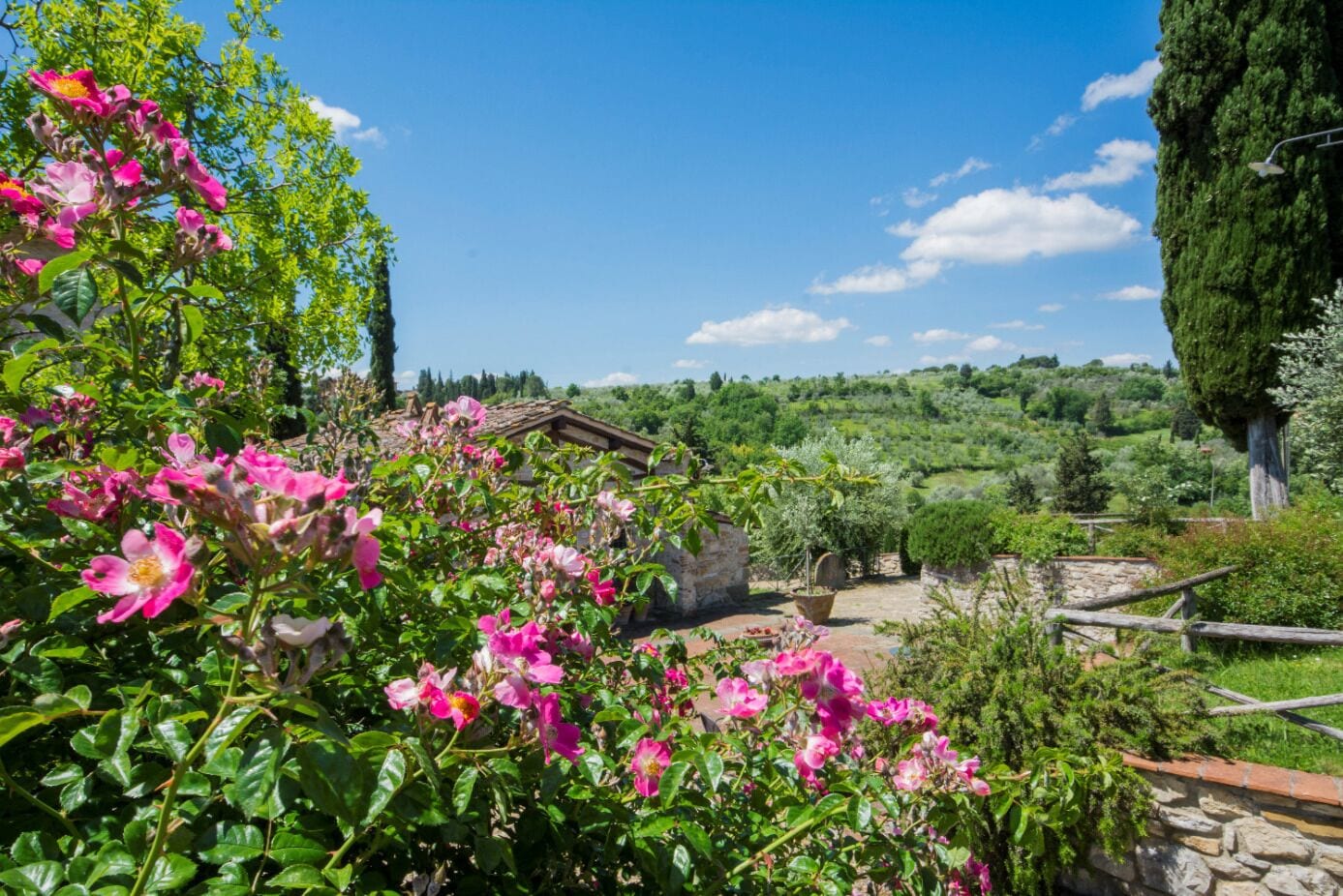 A rustic stone building with a garden in front, surrounded by lush greenery and hills in the background.