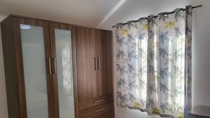 A bedroom with a wooden wardrobe, a bed with a floral duvet, and a window with a floral curtain. - Villa 1A on the complex of Casas del Sol  (Playa Blanca)