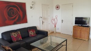 A living room with a black sofa, a glass coffee table, a television on a wooden cabinet, and a red rose painting on the wall. - Villa 1A on the complex of Casas del Sol  (Playa Blanca)