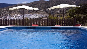 A swimming pool with two white umbrellas, a wooden fence, and a view of a town in the background.