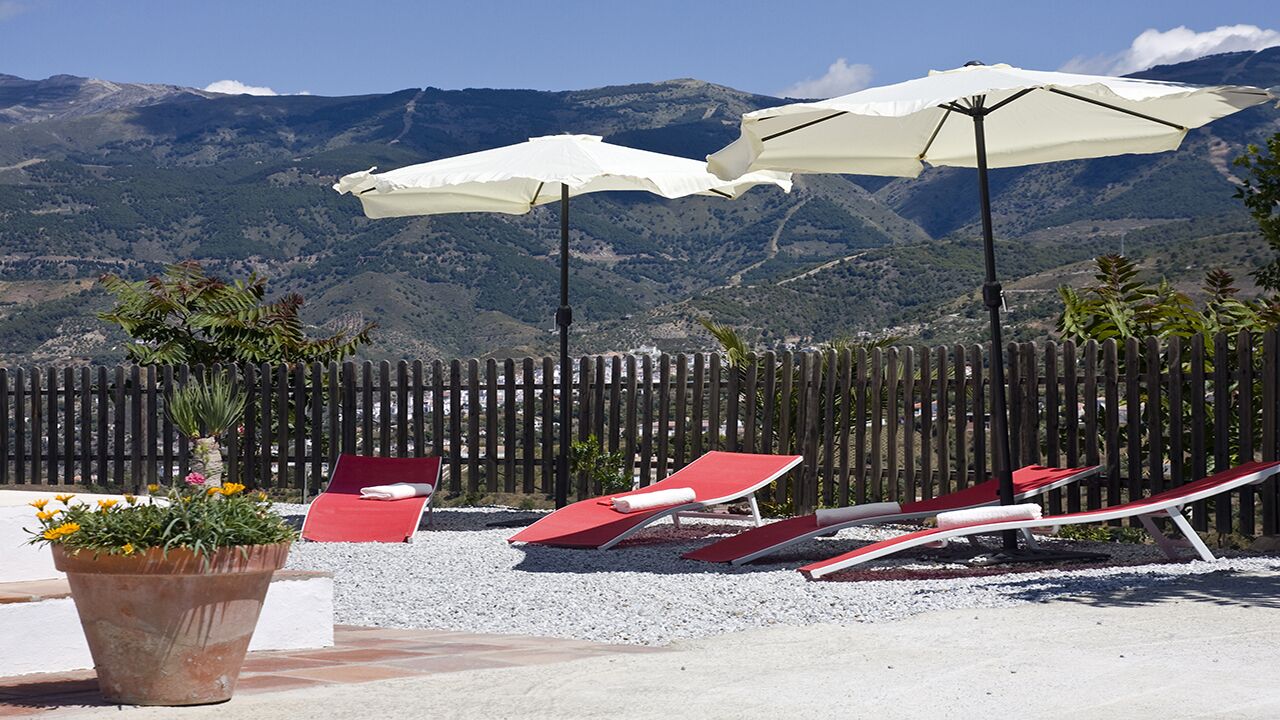 A patio with red lounge chairs and white umbrellas, a potted plant, and a view of mountains.