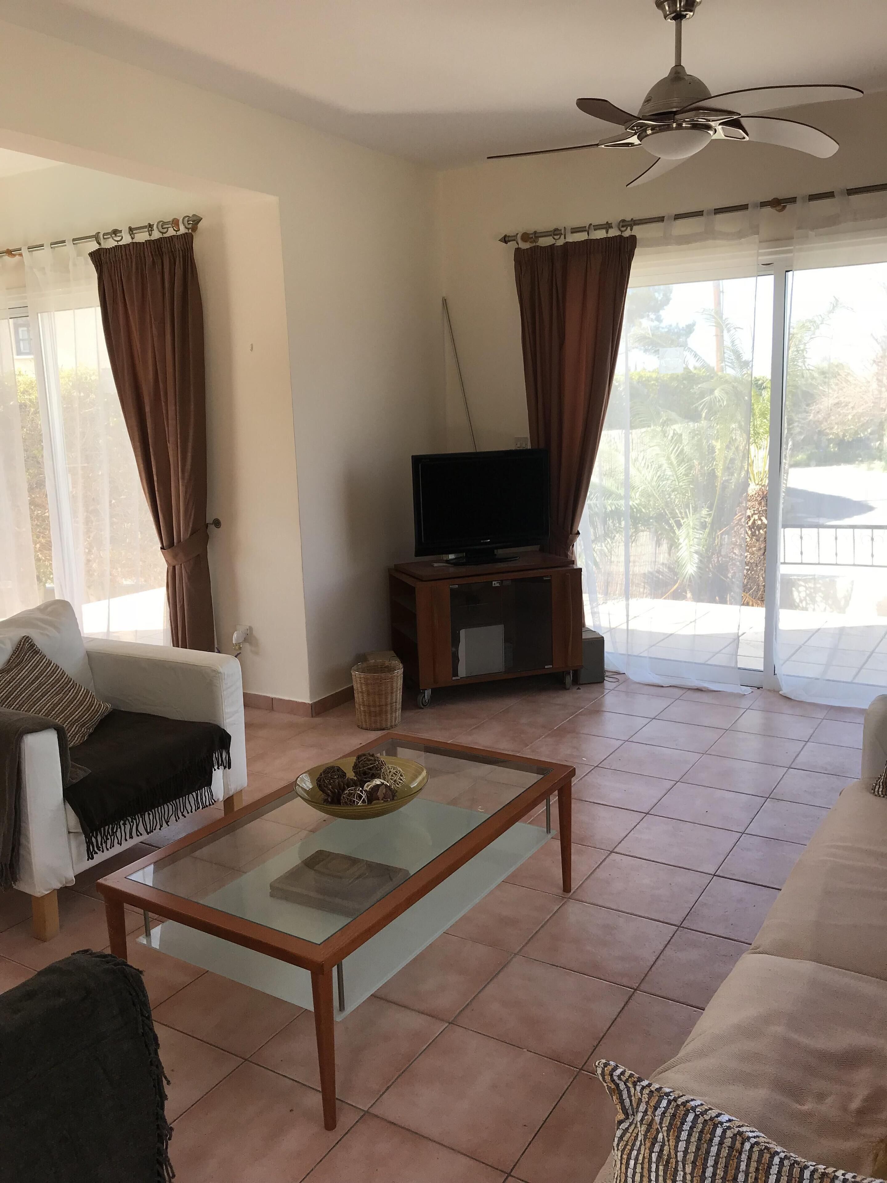 A living room with a glass coffee table, a sofa, a television on a wooden stand, and a ceiling fan.