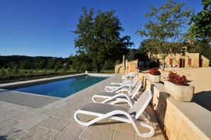 Zona junto a la piscina con sillones de descanso, piscina de piedra y un edificio con contraventanas rojas.