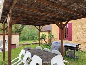 Un patio al aire libre con una mesa de madera y sillas blancas, una parrilla y un edificio de piedra al fondo.