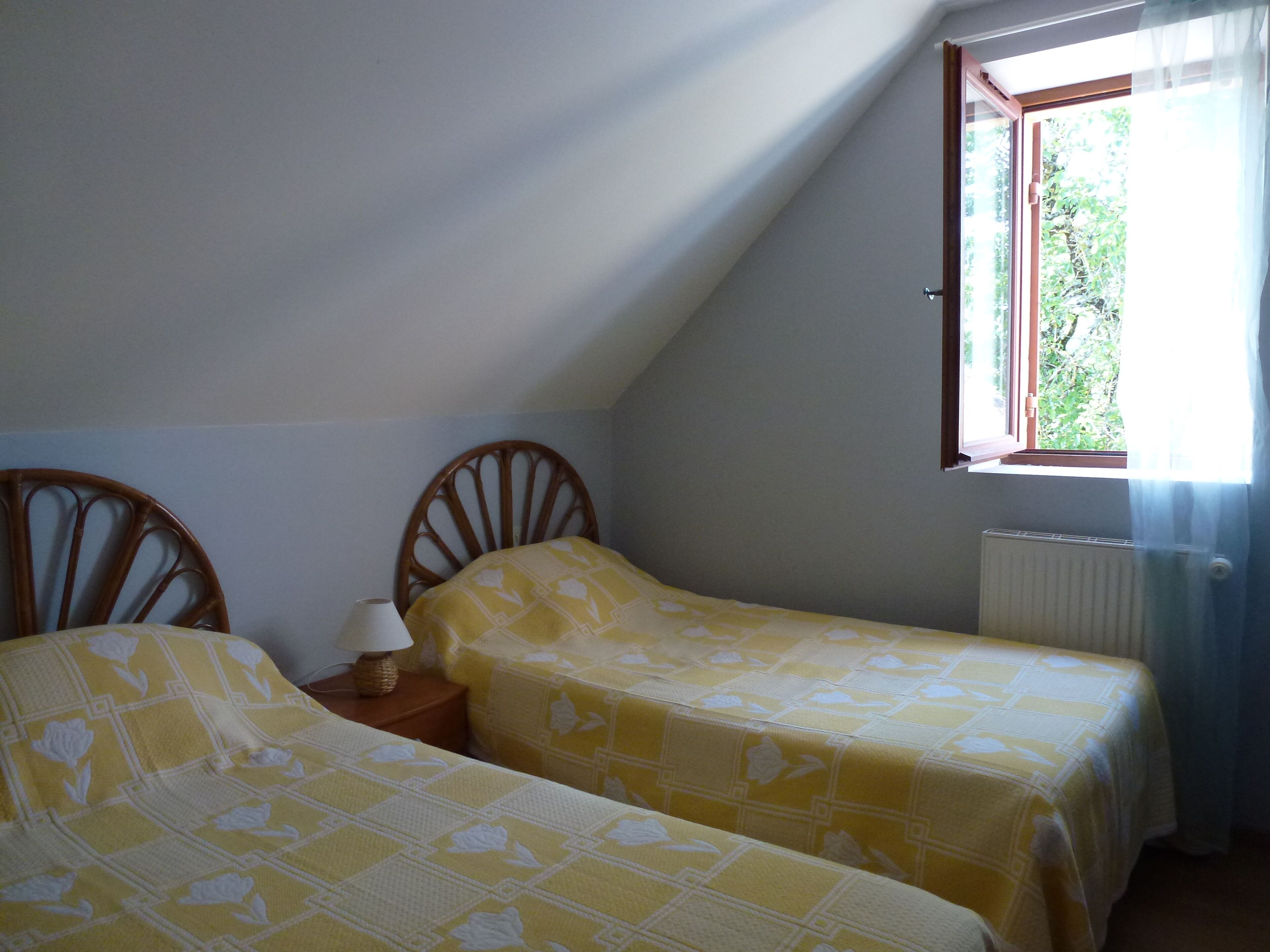 Une petite pièce faiblement éclairée, avec deux lits simples, une chaise en bois, une table de chevet avec une lampe et une fenêtre avec des rideaux.