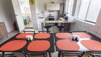 Una cocina moderna con armarios blancos, una estufa a gas y un microondas. Hay un área de comedor con mesas y sillas, y se ve la cocina a través de una puerta abierta.