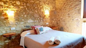 A room with a stone wall, a bed with white linens, two pillows, and two bedside tables. - Rural house in Girona. 11 bedrooms, up to 30 people, private pool. (Cabanelles)