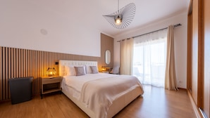 A modern hotel room with a large bed, wooden flooring, a nightstand with a lamp, a wall-mounted mirror, and a view of the outdoors through the window. - Spacious villa near town [46] (Lagos)