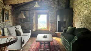 A rustic room with a stone wall, a fireplace, a sofa, a table, and a window with a view. - Etxetxobarri, a single-family farmhouse with views of the Anboto mountain range. (Mallabia - bizkaia)