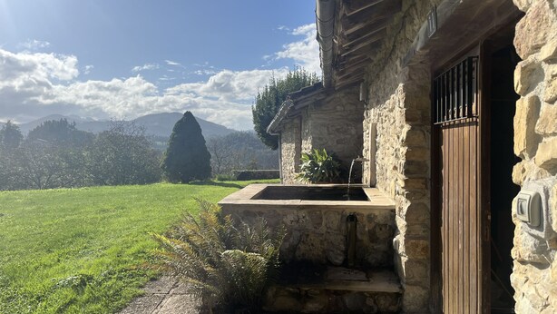 A stone building with a wooden door, a small garden, and a view of mountains in the background. - Etxetxobarri, a single-family farmhouse with views of the Anboto mountain range. (Mallabia - bizkaia)