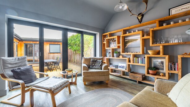 A spacious living area with a large sliding glass door leading to an outdoor patio, wooden shelving with books and decorative items, and comfortable seating. - The Old Butcher's Stores, Heacham, Norfolk (Heacham)