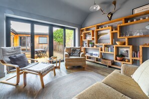 Un vaste espace de vie avec une grande porte-fenêtre coulissante donnant sur une terrasse extérieure, des étagères en bois ornées de livres et d’objets décoratifs, et des sièges confortables.