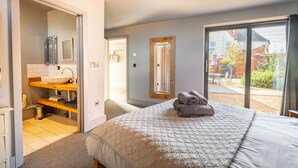 A bedroom with a large bed, a bathroom with a sink and shelves, and a view of a garden through a sliding door. - The Old Butcher's Stores, Heacham, Norfolk (Heacham)