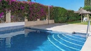 Una piscina con agua azul cristalina, rodeada por un muro blanco y un seto de flores moradas.