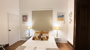 A hotel room with a bed, two bedside tables, a window with blinds, and artwork on the walls.