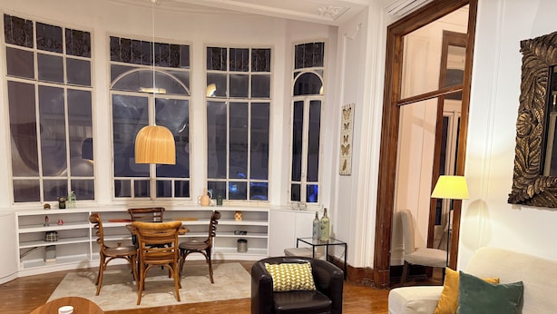 A cozy living room with a dining area, wooden furniture, and large windows with decorative glass panes.
