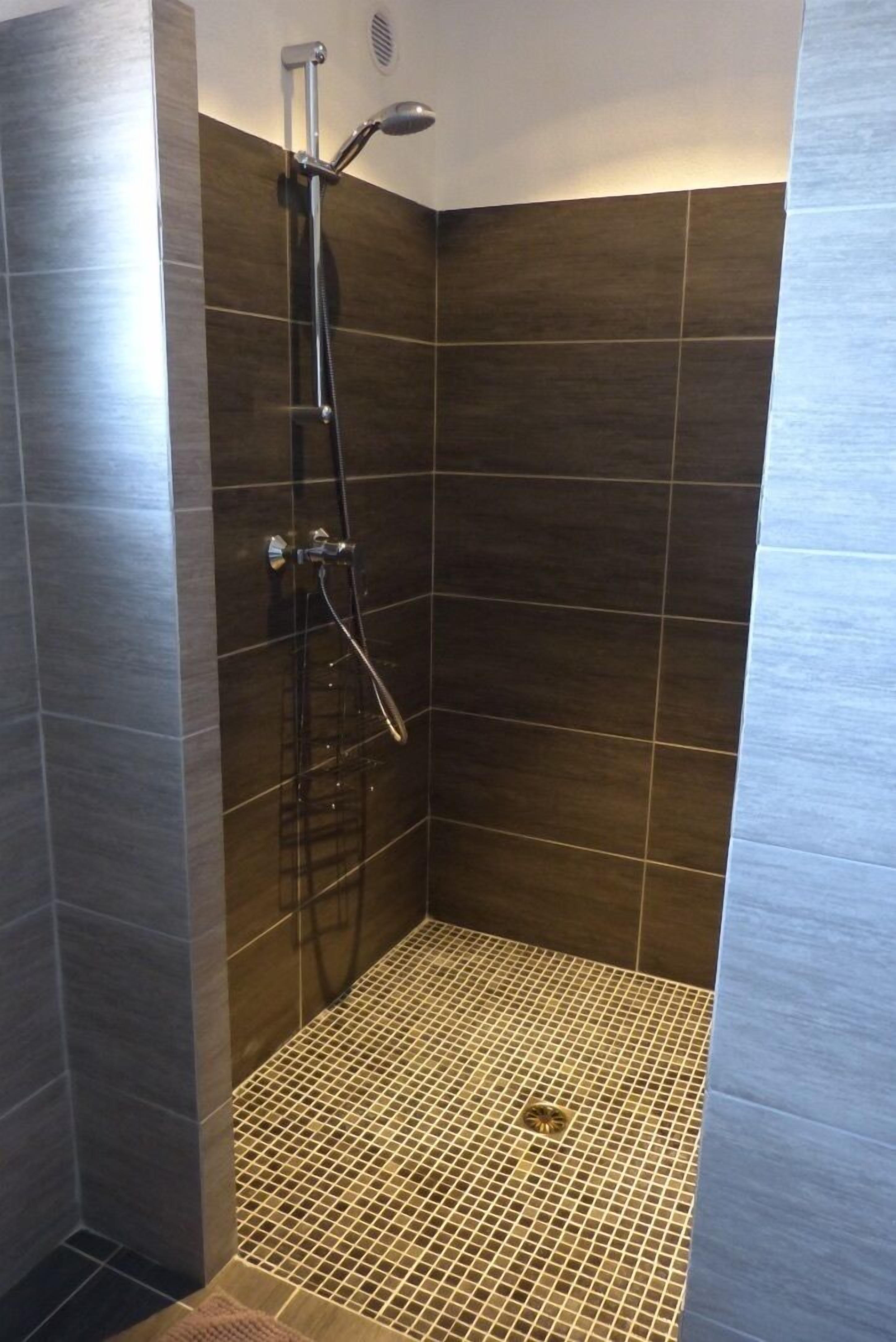 A shower with a handheld showerhead and a wall-mounted faucet, tiled walls, and a floor with a drainage grate.