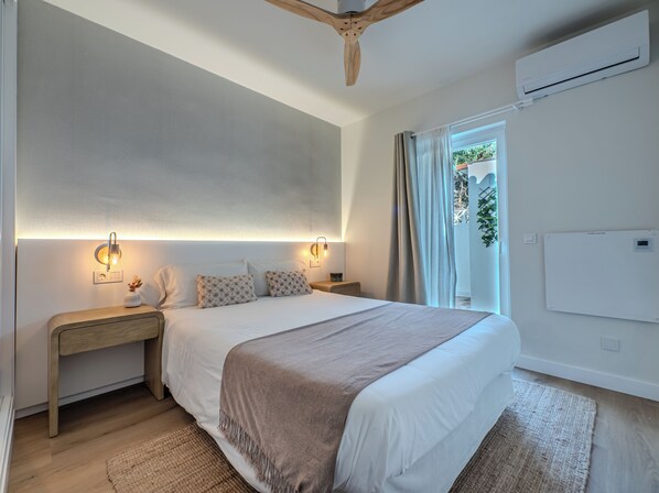 A modern bedroom with a large bed, bedside tables, a ceiling fan, and a view of greenery outside.