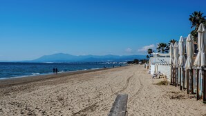 A beach with a wooden walkway leading to the sea, umbrellas, and a mountain in the background. - House 80 m from the beach, garden, swimming pool, barbecue and Wifi Ideal for families. (Marbella)