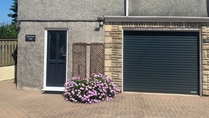 Una casa a due piani con facciata in pietra, un garage con basculante e un piccolo giardino con fiori rosa.