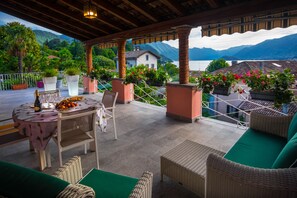 Une terrasse couverte, meublée de meubles en osier, avec une table sur laquelle se trouve un panier de fruits et des plantes en pot.
