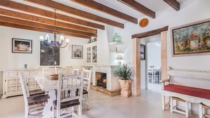 A spacious room with a dining area featuring a white table and chairs, a fireplace, and a bench with a red cushion. - Villa Can Boseta by SunVillas Mallorca (Pollença)