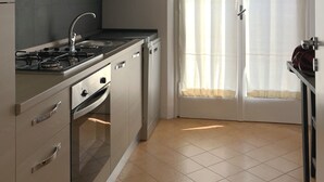Une cuisine moderne équipée d’une cuisinière à gaz, d’un four et d’un lave-vaisselle. Il y a une fenêtre avec des rideaux légers et une porte dotée d’une poignée.