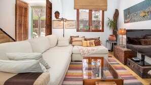 Un salon spacieux avec un grand canapé d’angle, un téléviseur et une table basse. Le plafond est en bois, avec des poutres apparentes, et une grande fenêtre offre une vue dégagée. Une porte donne accès à l’extérieur.