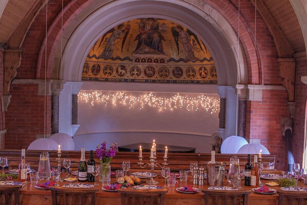 Une salle de banquet dotée d’un haut plafond en arc, d’une fresque murale et d’une longue table dressée pour un événement officiel.