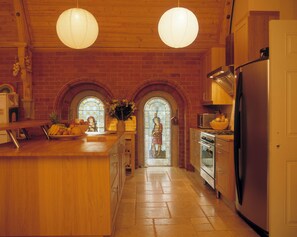 A kitchen with wooden cabinets, a stainless steel refrigerator, a stove, and a large island with a bowl of fruit. There are two hanging paper lanterns and a window with a stained glass design.