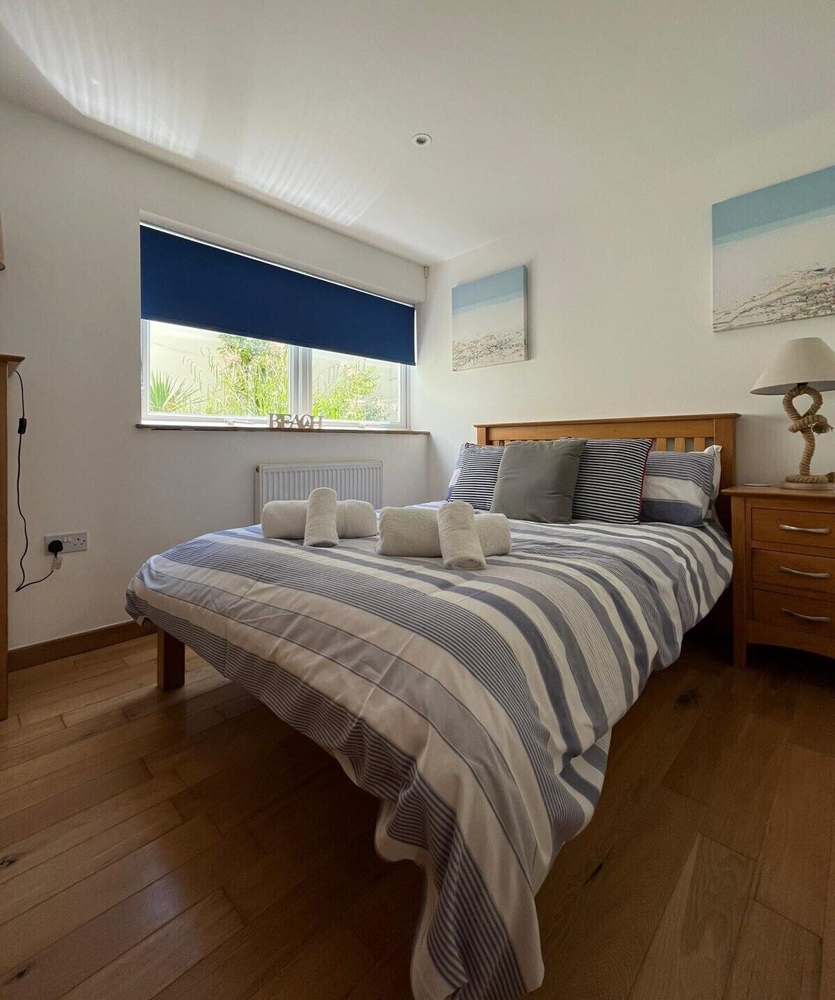 A bedroom with a large bed, a window with a blue blind, a bedside table with a lamp, and a radiator under the window.